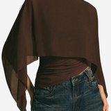 A woman wearing a dark brown draped long-sleeve top with a wide boat neckline and sheer side panels, paired with blue jeans, against a plain light background.