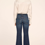 A woman shown from the back wearing a white long-sleeve button-up shirt and dark blue flared denim jeans, paired with black heels, against a plain light beige studio background.