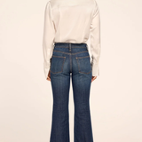 A woman shown from the back wearing a white long-sleeve button-up shirt and dark blue flared denim jeans, paired with black heels, against a plain light beige studio background.