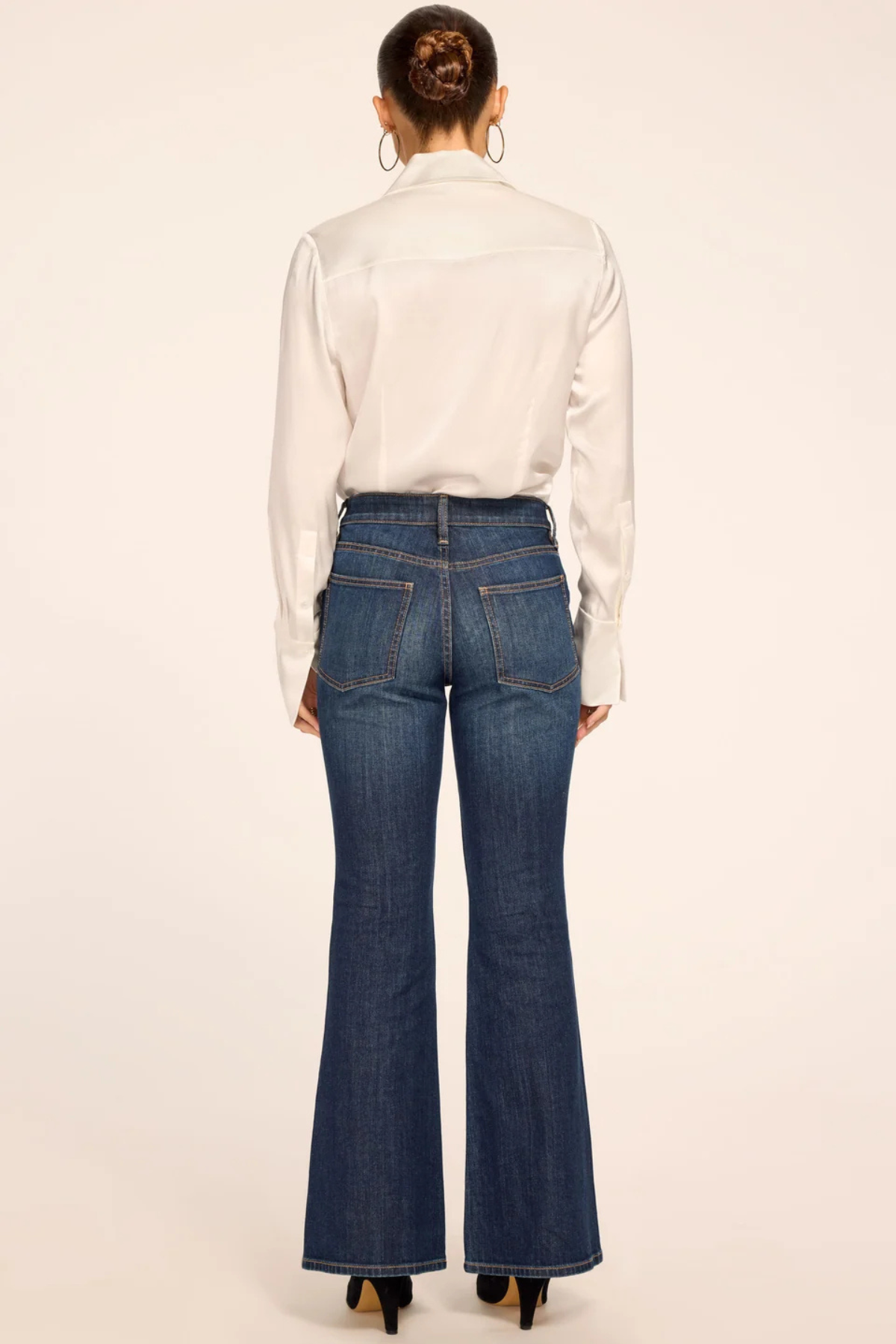 A woman shown from the back wearing a white long-sleeve button-up shirt and dark blue flared denim jeans, paired with black heels, against a plain light beige studio background.