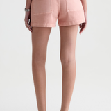 A person shown from the back wearing light pink denim shorts, a gray top, and dark brown sandals against a plain light gray studio background.