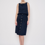 A model stands against a light gray studio background wearing a sleeveless dark navy dress with a studded neckline and a front button placket. She is also wearing beige high-heeled sandals and hoop earrings.