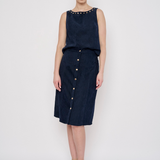 A model stands against a light gray studio background wearing a sleeveless dark navy dress with a studded neckline and a front button placket. She is also wearing beige high-heeled sandals and hoop earrings.