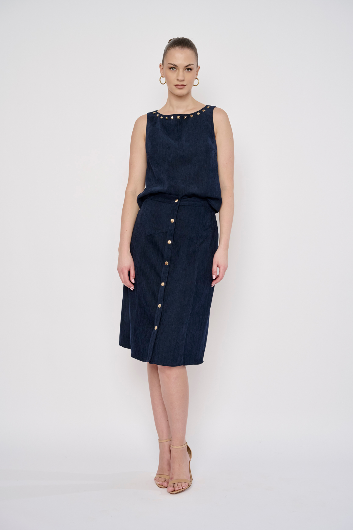 A model stands against a light gray studio background wearing a sleeveless dark navy dress with a studded neckline and a front button placket. She is also wearing beige high-heeled sandals and hoop earrings.