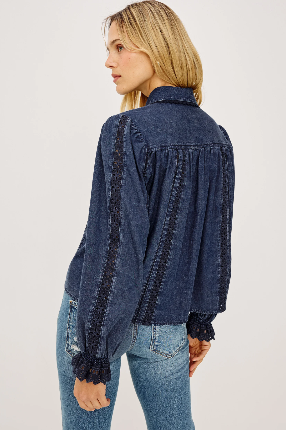 A woman shown from the back wearing a dark blue denim long-sleeve blouse with black lace trim along the shoulders, sleeves, and cuffs, paired with light blue jeans against a plain white background.