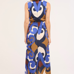 A woman viewed from the back wearing a sleeveless, high-waisted maxi dress with a bold abstract swirl pattern in blue, brown, cream, and white, paired with gray heels, against a plain white studio background.