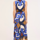 A woman viewed from the back wearing a sleeveless, high-waisted maxi dress with a bold abstract swirl pattern in blue, brown, cream, and white, paired with gray heels, against a plain white studio background.