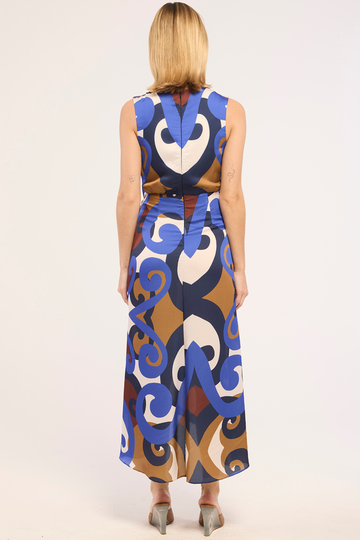 A woman viewed from the back wearing a sleeveless, high-waisted maxi dress with a bold abstract swirl pattern in blue, brown, cream, and white, paired with gray heels, against a plain white studio background.