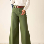 A person wearing olive green wide-leg pants with a brown belt, styled with a white long-sleeve top and brown sandals, standing against a plain beige wall.