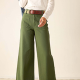 A person wearing olive green wide-leg pants with a brown belt, styled with a white long-sleeve top and brown sandals, standing against a plain beige wall.