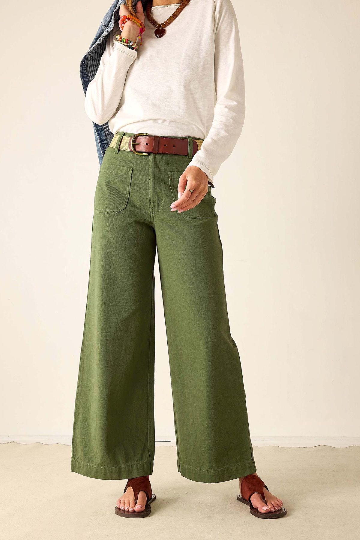 A person wearing olive green wide-leg pants with a brown belt, styled with a white long-sleeve top and brown sandals, standing against a plain beige wall.
