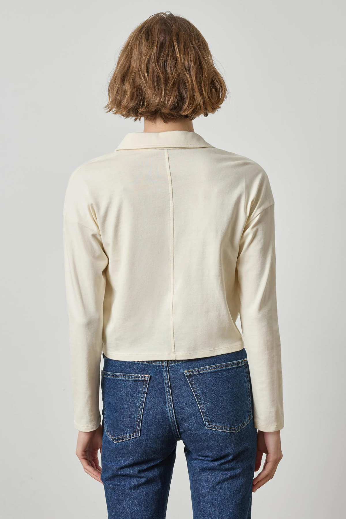 A model viewed from the back wearing a cream-colored long-sleeve knit top and blue jeans against a light gray studio background.