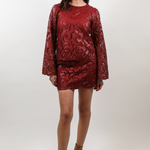 A woman modeling a matching red long-sleeve top and mini skirt set with a textured floral pattern, paired with brown suede ankle boots, against a plain light gray studio background.