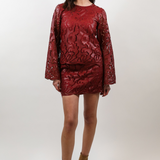 A woman modeling a matching red long-sleeve top and mini skirt set with a textured floral pattern, paired with brown suede ankle boots, against a plain light gray studio background.