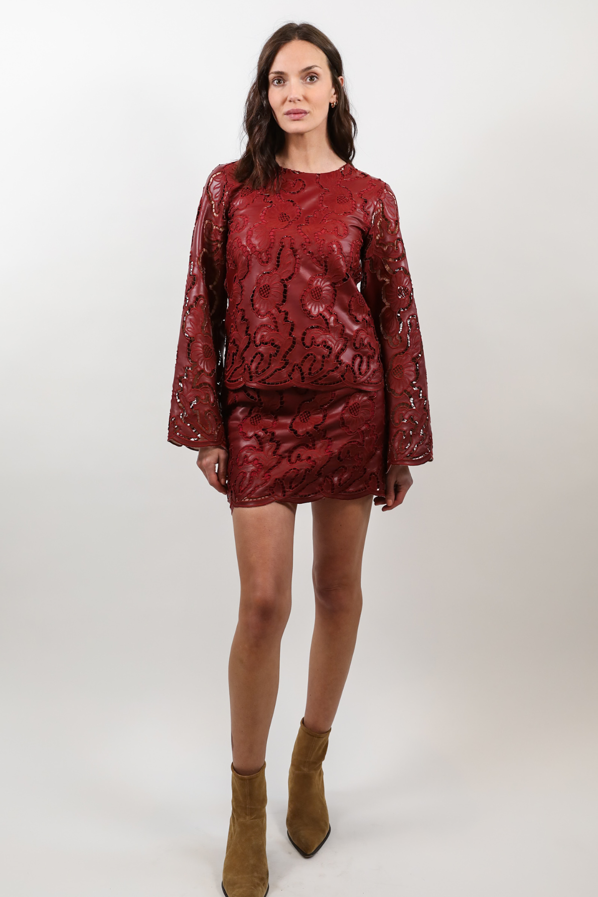 A woman modeling a matching red long-sleeve top and mini skirt set with a textured floral pattern, paired with brown suede ankle boots, against a plain light gray studio background.
