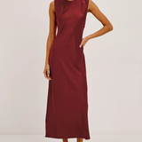 A woman modeling a sleeveless, high-neck midi dress in a deep burgundy or maroon satin-like fabric, paired with black strappy high-heeled sandals, against a plain off-white studio background.