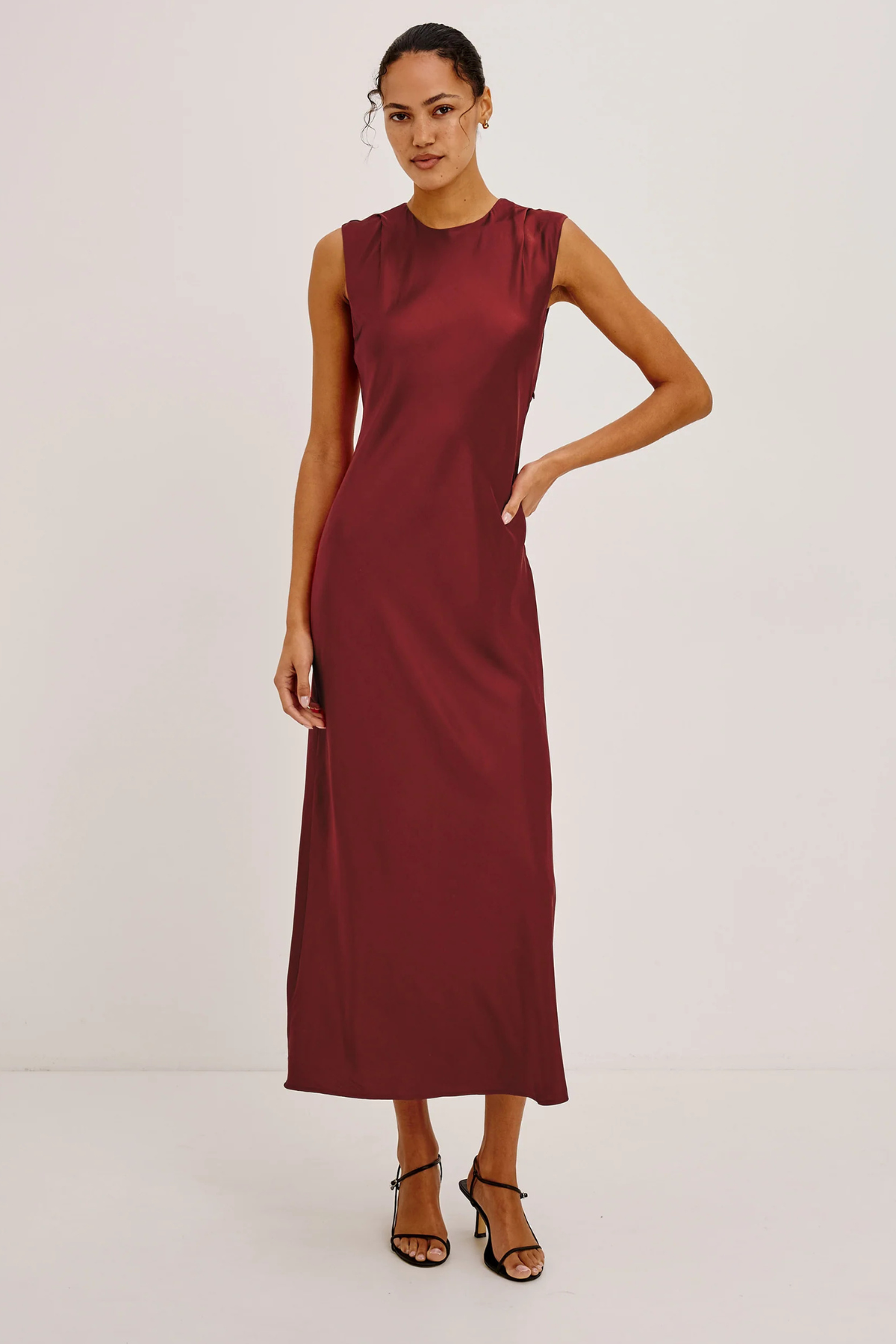 A woman modeling a sleeveless, high-neck midi dress in a deep burgundy or maroon satin-like fabric, paired with black strappy high-heeled sandals, against a plain off-white studio background.