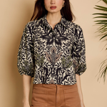 A woman modeling a black and beige botanical print blouse with three-quarter sleeves, paired with brown trousers, set against a warm beige background with a leafy plant on the side.
