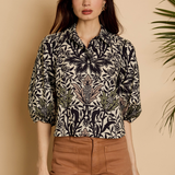 A woman modeling a black and beige botanical print blouse with three-quarter sleeves, paired with brown trousers, set against a warm beige background with a leafy plant on the side.