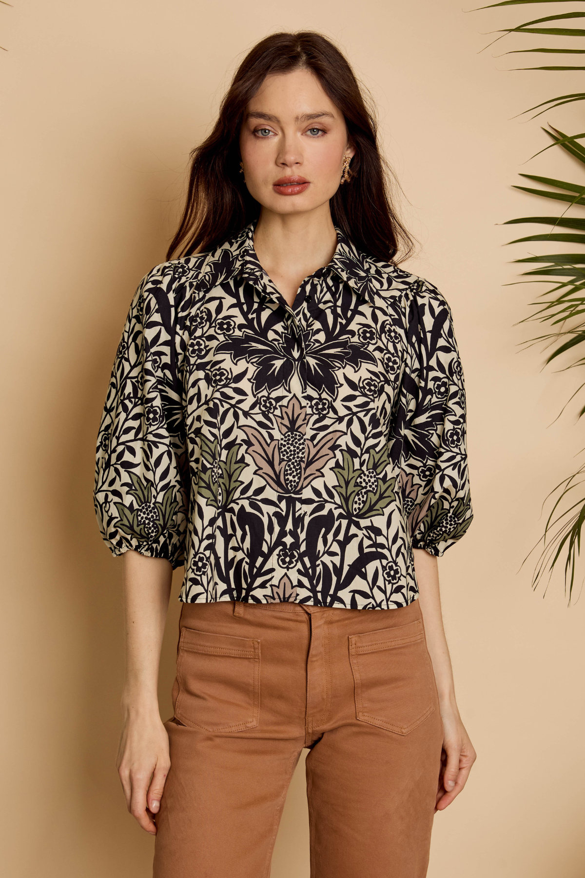 A woman modeling a black and beige botanical print blouse with three-quarter sleeves, paired with brown trousers, set against a warm beige background with a leafy plant on the side.