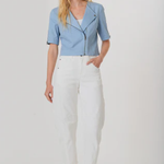 A woman modeling a light blue short-sleeve moto jacket with a front zipper, paired with white cropped pants and brown woven sandals, against a plain light gray studio background.