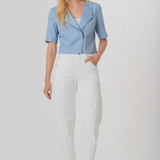 A woman modeling a light blue short-sleeve moto jacket with a front zipper, paired with white cropped pants and brown woven sandals, against a plain light gray studio background.