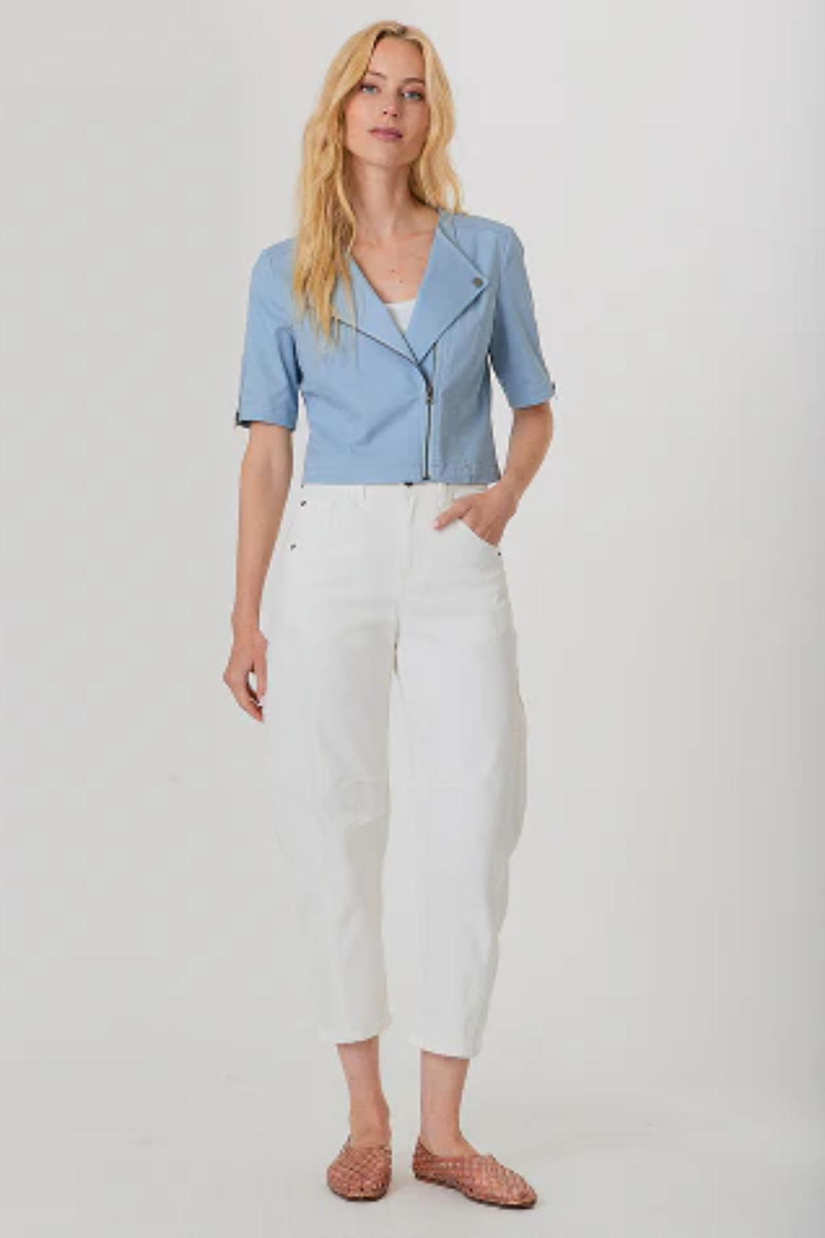A woman modeling a light blue short-sleeve moto jacket with a front zipper, paired with white cropped pants and brown woven sandals, against a plain light gray studio background.