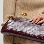 A close-up of a person holding a dark burgundy crocodile-embossed clutch bag with a gold-tone metal frame and clasp.