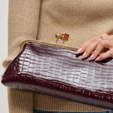 A close-up of a person holding a dark burgundy crocodile-embossed clutch bag with a gold-tone metal frame and clasp.