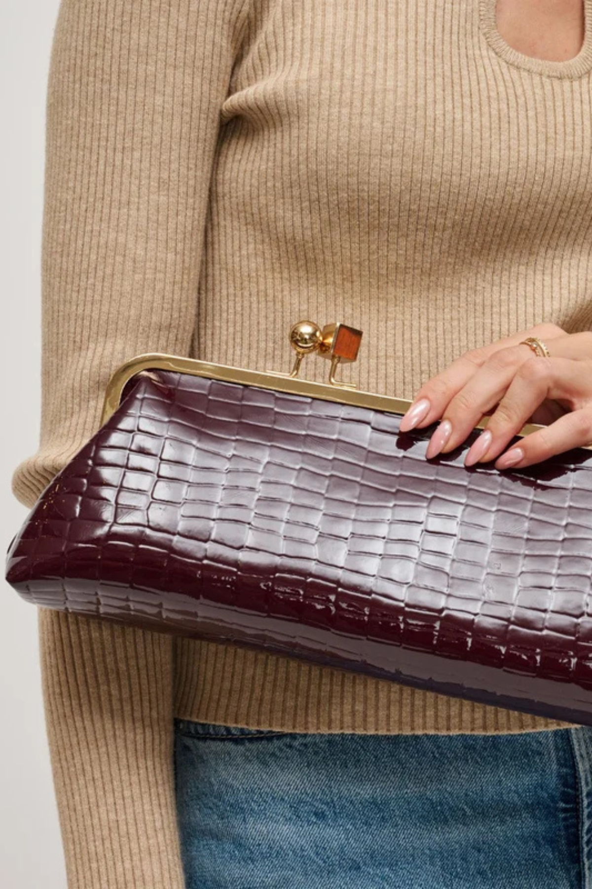 A close-up of a person holding a dark burgundy crocodile-embossed clutch bag with a gold-tone metal frame and clasp.