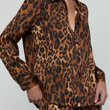 A model wearing a matching leopard-print long-sleeve button-up shirt and trousers set, shown from the neck down against a light gray studio background.