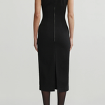 A model shown from the back wearing a sleeveless black midi dress with a front slit and a central zipper, paired with black tights and black heels, against a light gray studio background.