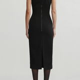 A model shown from the back wearing a sleeveless black midi dress with a front slit and a central zipper, paired with black tights and black heels, against a light gray studio background.