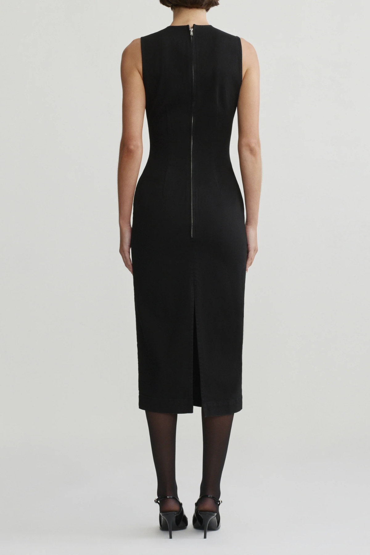 A model shown from the back wearing a sleeveless black midi dress with a front slit and a central zipper, paired with black tights and black heels, against a light gray studio background.