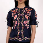 A model wearing a navy blue short-sleeve blouse with pink floral embroidery and tie details on the sleeves, paired with black trousers, against a plain white background.
