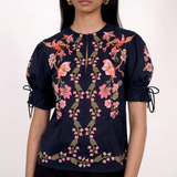 A model wearing a navy blue short-sleeve blouse with pink floral embroidery and tie details on the sleeves, paired with black trousers, against a plain white background.