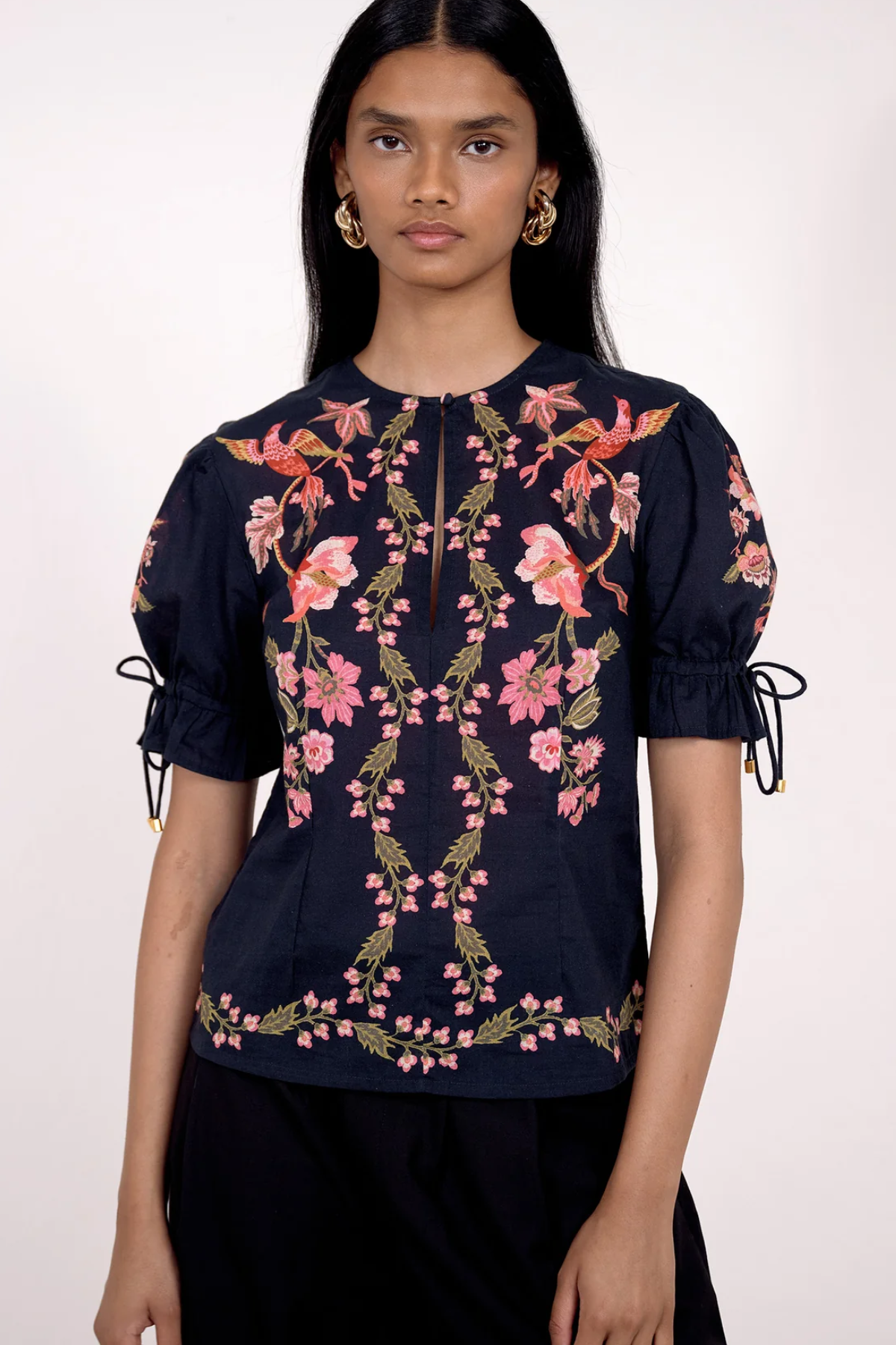 A model wearing a navy blue short-sleeve blouse with pink floral embroidery and tie details on the sleeves, paired with black trousers, against a plain white background.