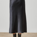 A black satin midi skirt worn by a model, shown from the back against a neutral studio background with a concrete floor.
