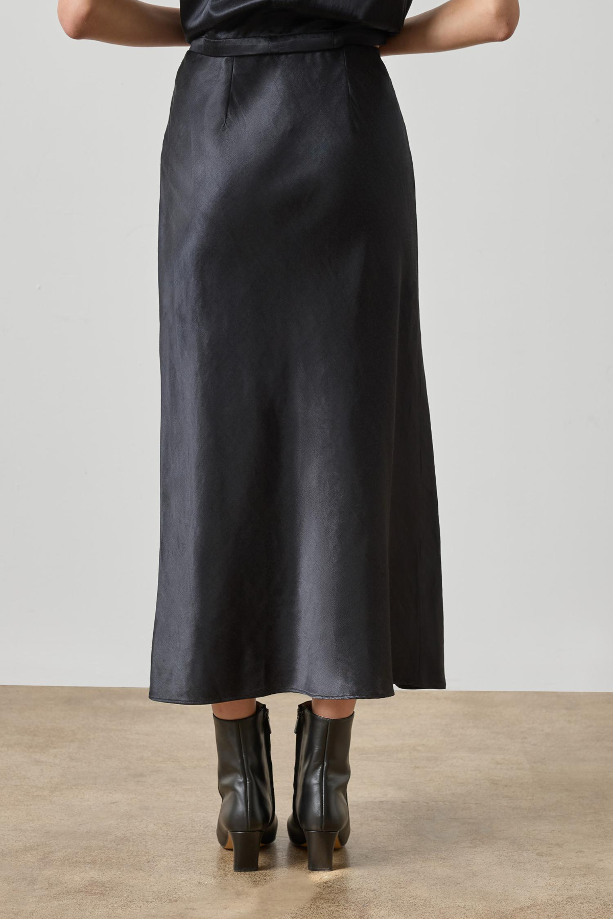 A black satin midi skirt worn by a model, shown from the back against a neutral studio background with a concrete floor.