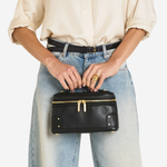 A person wearing a cream blouse, black belt, and light-wash wide-leg jeans holds a small black leather cosmetic case with gold-tone hardware against a plain light background.