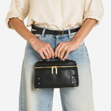 A person wearing a cream blouse, black belt, and light-wash wide-leg jeans holds a small black leather cosmetic case with gold-tone hardware against a plain light background.