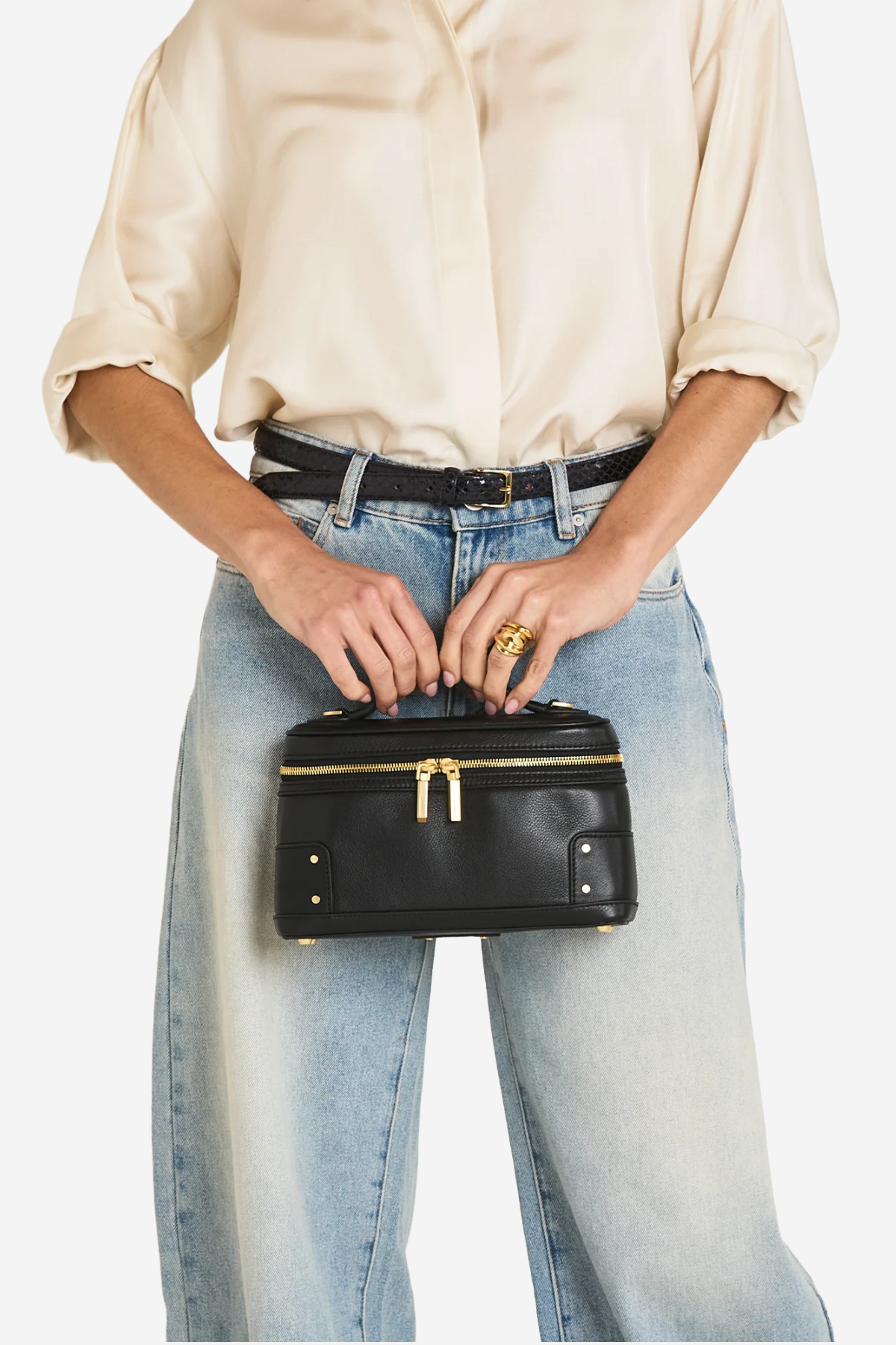 A person wearing a cream blouse, black belt, and light-wash wide-leg jeans holds a small black leather cosmetic case with gold-tone hardware against a plain light background.