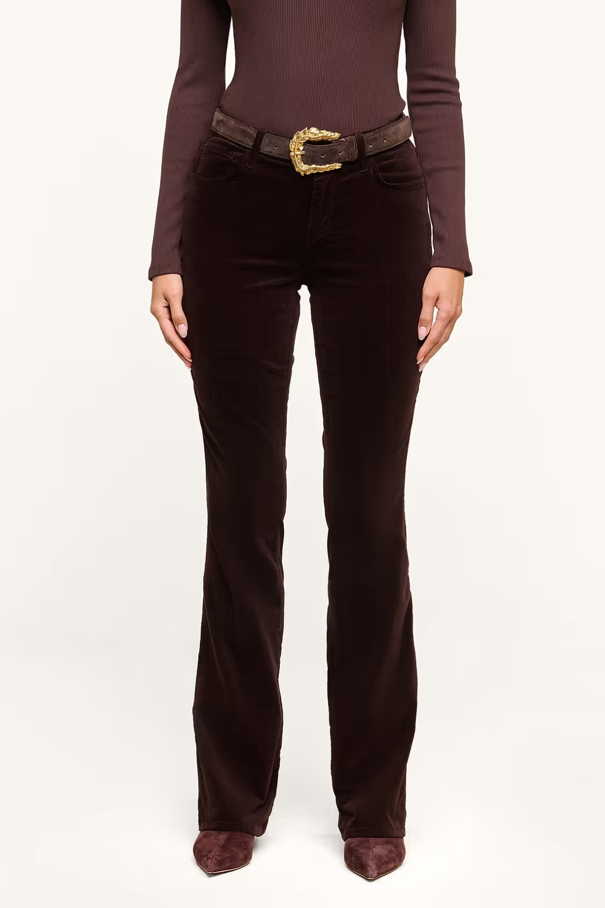A model wearing dark brown velvet flare pants with a dark brown belt featuring a gold buckle, paired with a matching dark brown long-sleeve top and pointed-toe heels, shot against a plain white studio background.