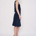 A woman stands in profile wearing a sleeveless dark navy midi dress with a textured fabric and nude high-heeled sandals against a plain white studio background.