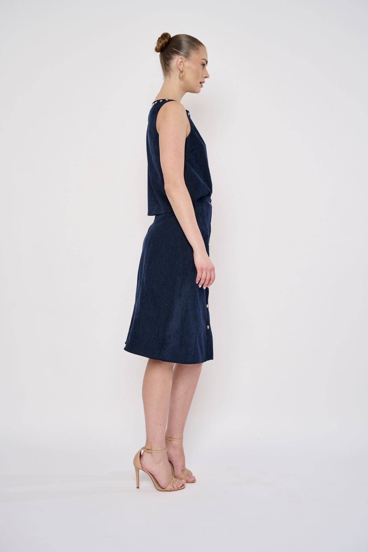 A woman stands in profile wearing a sleeveless dark navy midi dress with a textured fabric and nude high-heeled sandals against a plain white studio background.