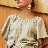 Woman wearing a green outfit with sunglasses and earrings against a textured wall.