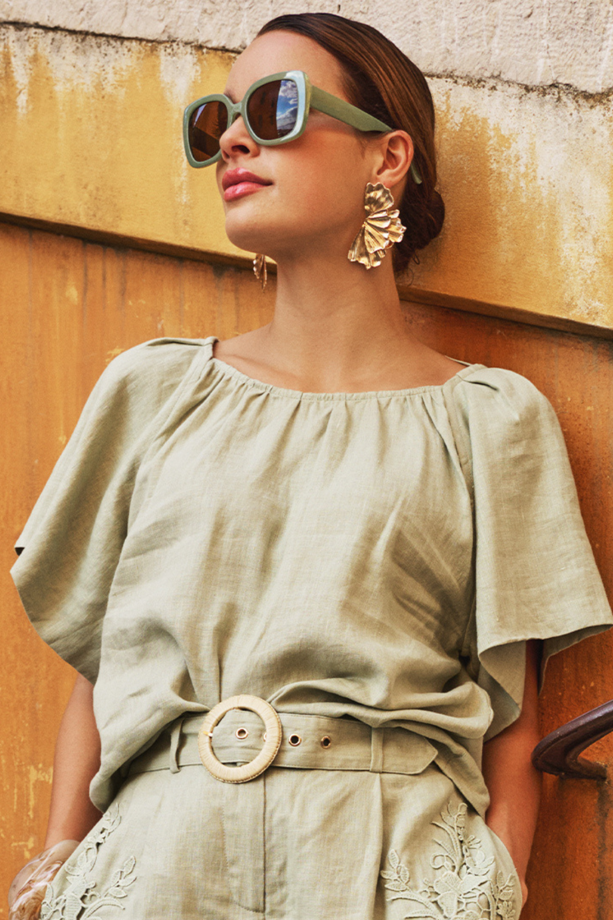 Woman wearing a green outfit with sunglasses and earrings against a textured wall.