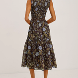 A woman modeling a sleeveless midi dress with a ruffled neckline and a tiered skirt, shown from the back. The dress features a dark brown base with a blue and beige floral pattern.