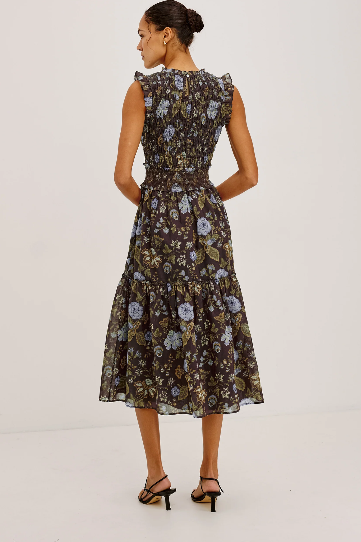 A woman modeling a sleeveless midi dress with a ruffled neckline and a tiered skirt, shown from the back. The dress features a dark brown base with a blue and beige floral pattern.