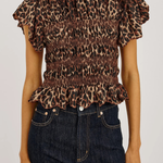 A woman wearing a brown leopard-print smocked blouse with ruffled sleeves and a ruffled hem, paired with dark blue jeans, against a plain light background.